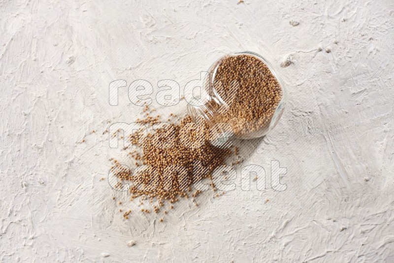 A glass spice jar full of mustard seeds and jar is flipped with fallen seeds on a textured white flooring