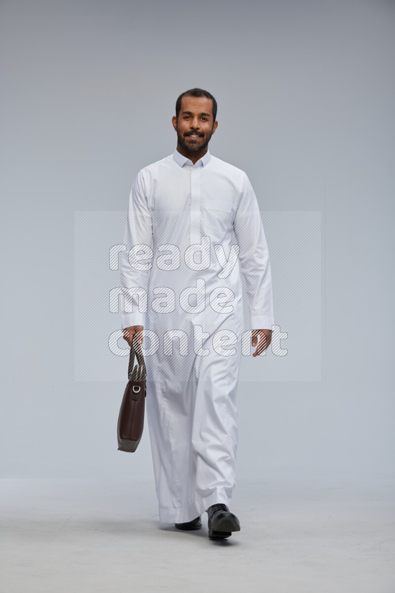 Saudi man wearing thob standing holding suitcase on gray background