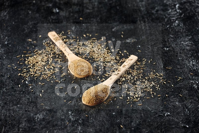 2 wooden spoons full of cumin powder with spreaded seeds on textured black flooring