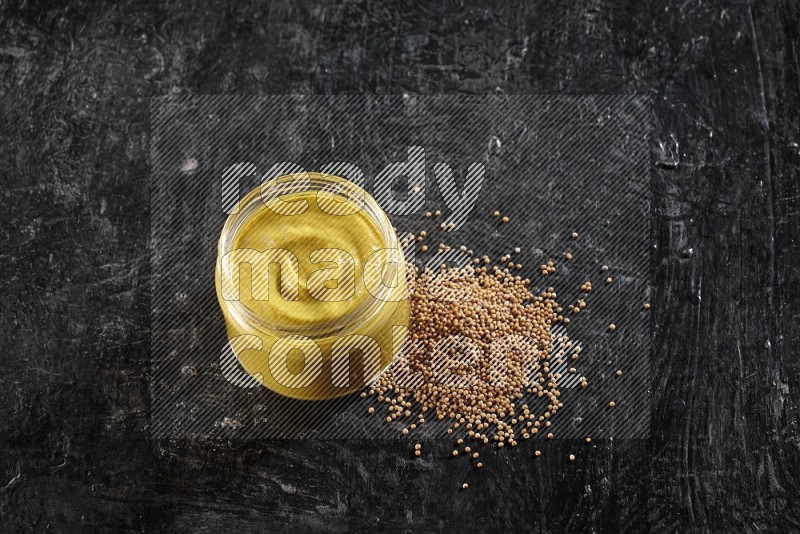 A glass jar full of mustard paste and mustard seeds spread next to it on a textured black flooring
