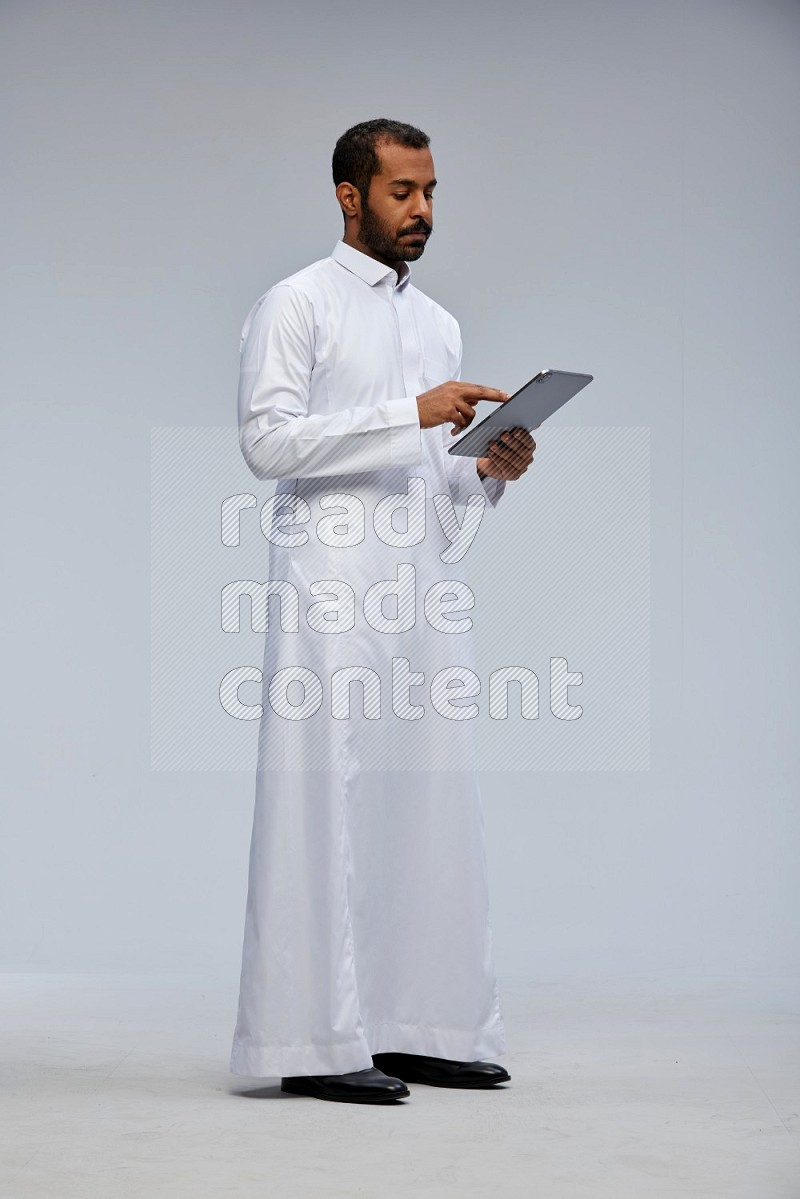 Saudi man wearing Thob standing working on tablet on Gray background