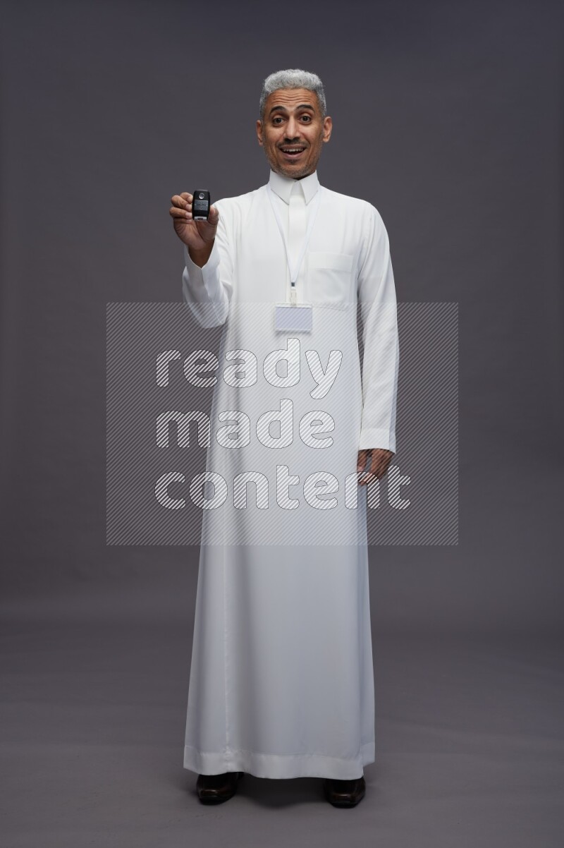 Saudi man wearing thob with neck strap employee badge standing holding key car on gray background