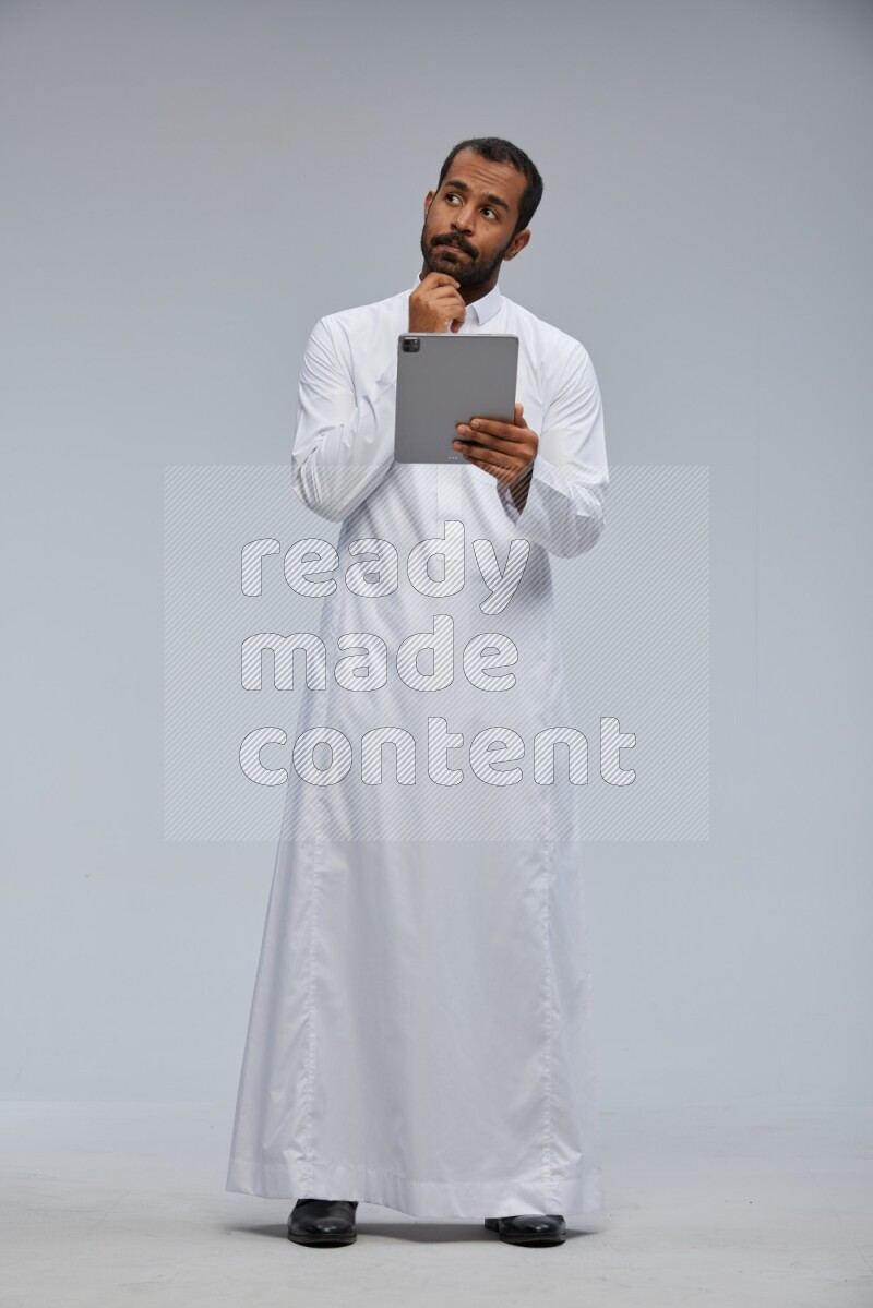 Saudi man wearing Thob standing working on tablet on Gray background