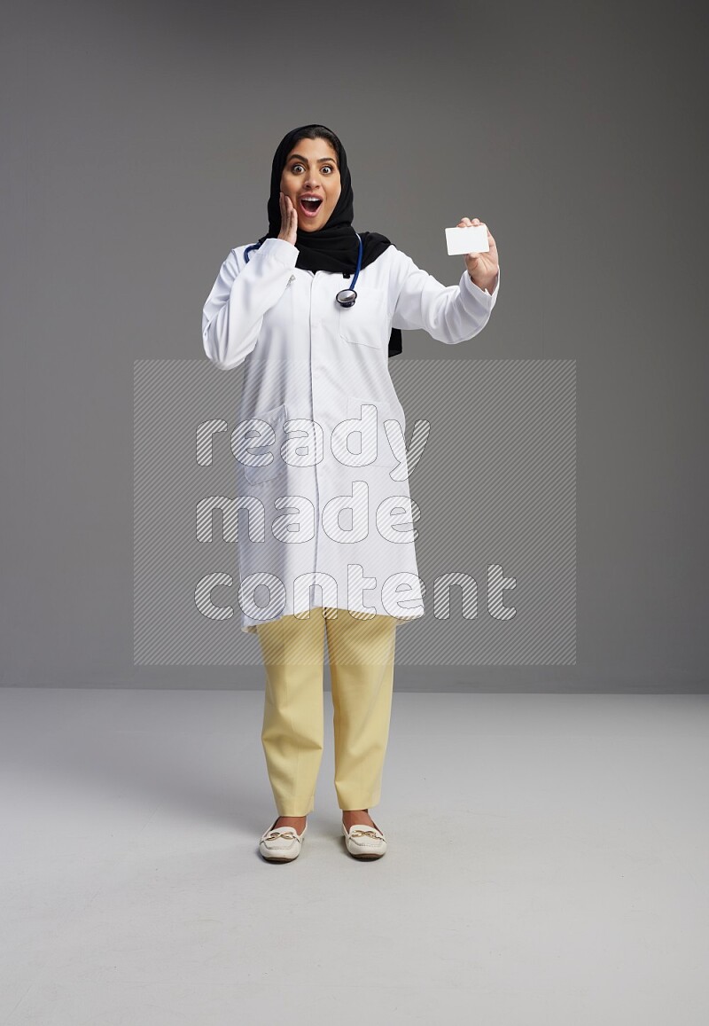 Saudi woman wearing lab coat with stethoscope standing holding ATM card on Gray background