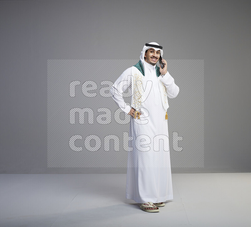 A saudi man standing wearing thob and white shomag with flag scarf on his neck talking on phone on gray background