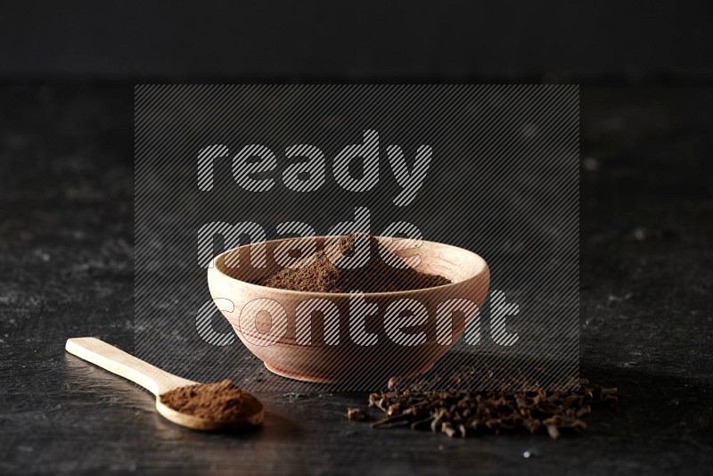 A wooden bowl and a wooden spoon full of cloves powder with spreaded cloves on a textured black flooring