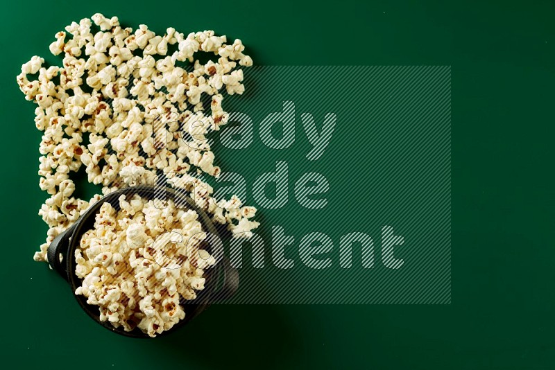 A black ceramic bowl full of popcorn with popcorn beside it on a green background in a top view shot