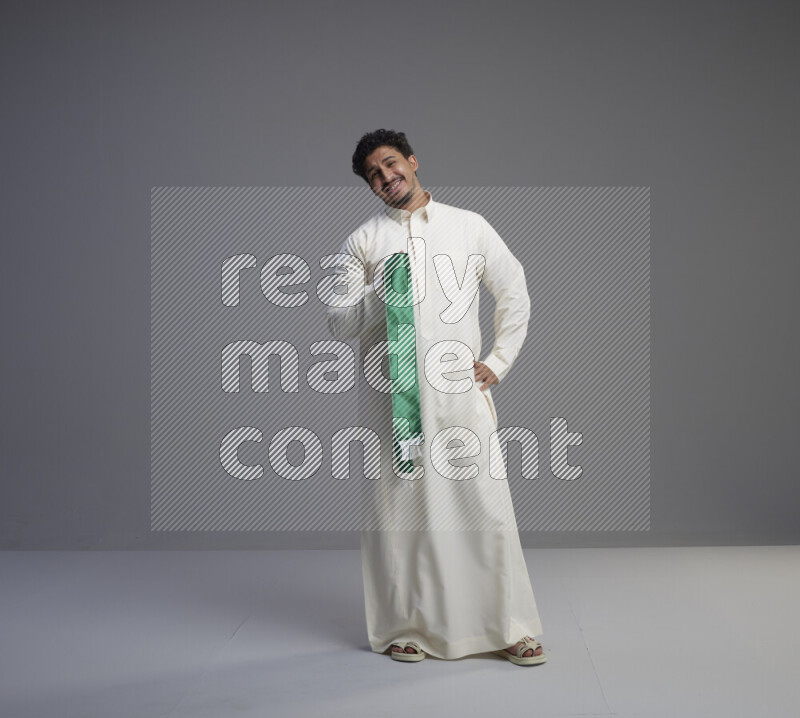 A Saudi man standing wearing thob holding Saudi flag scarf on gray background