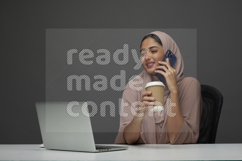 A Saudi woman Setting on her desk
 calling  on a Gray Background wearing Brown Abaya with Hijab