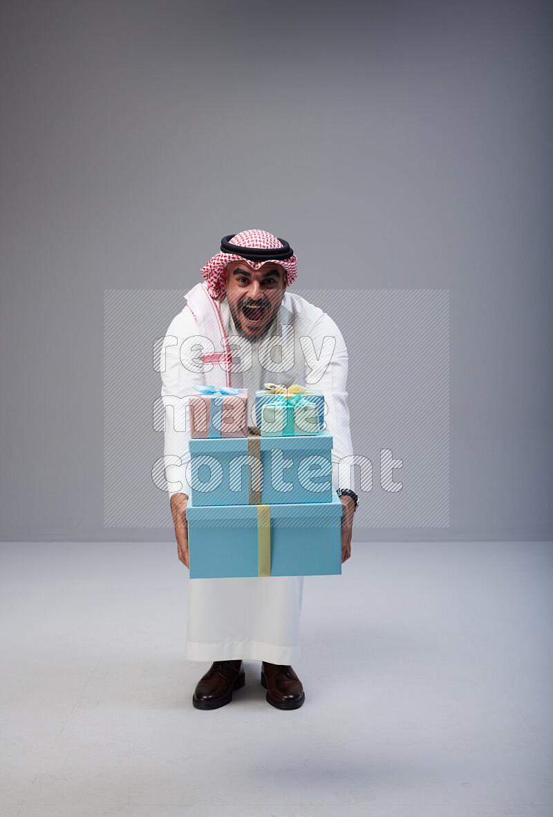 Saudi man Wearing Thob and red Shomag standing holding gift box on Gray background