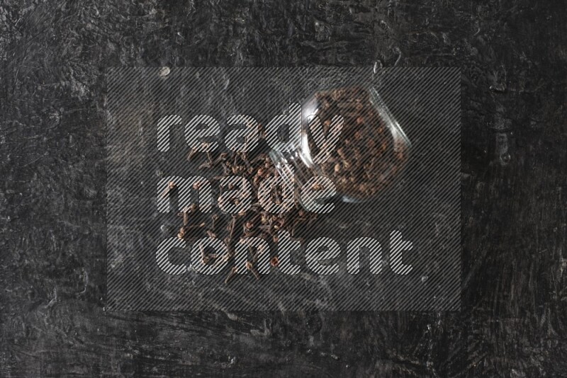 A flipped glass spice jar full of cloves and cloves came out of it on textured black flooring