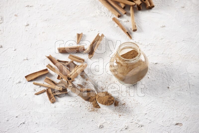Herbal glass jar full cinnamon powder and a metal spoon surrounded by cinnamon sticks on a white background