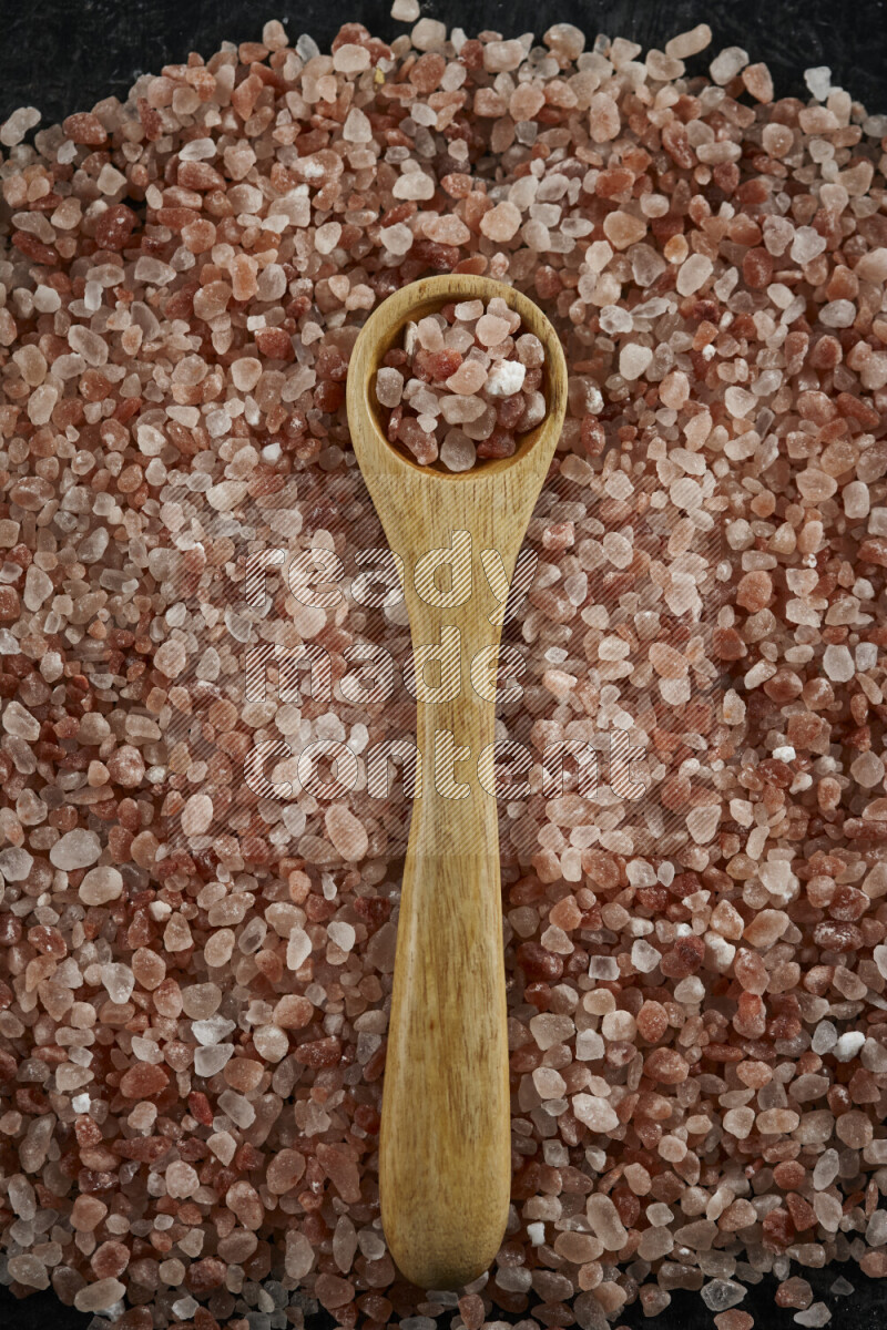 A wooden spoon full of coarse himalayan salt crystals on a bunch of the crystals on black background