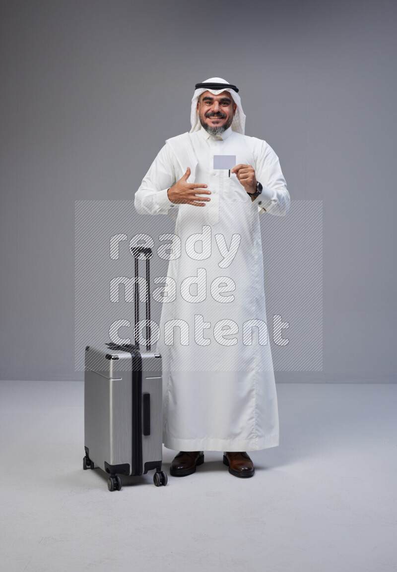 Saudi man wearing Thob and white Shomag standing holding Travel bag and ATM card on Gray background