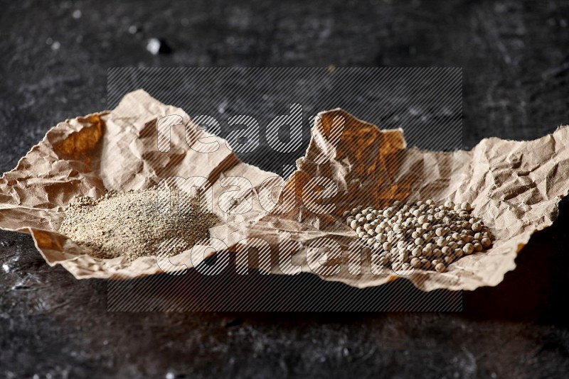 White pepper beads and powder in 2 crumpled paper on textured black flooring