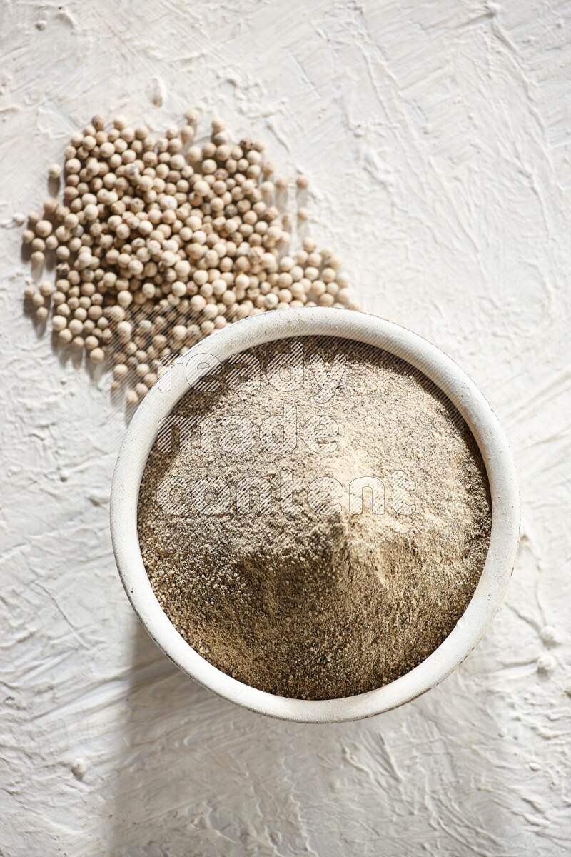 وعاء فخاري أبيض ممتلئ ببودرة الفلفل الأبيض مع حبوب الفلفل على أرضية بيضاء