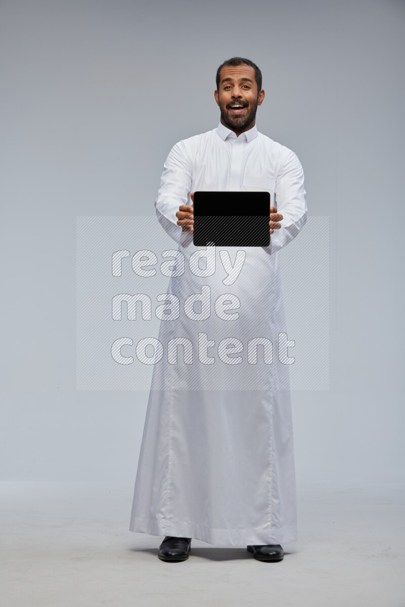 Saudi man wearing Thob standing showing tablet to camera on Gray background