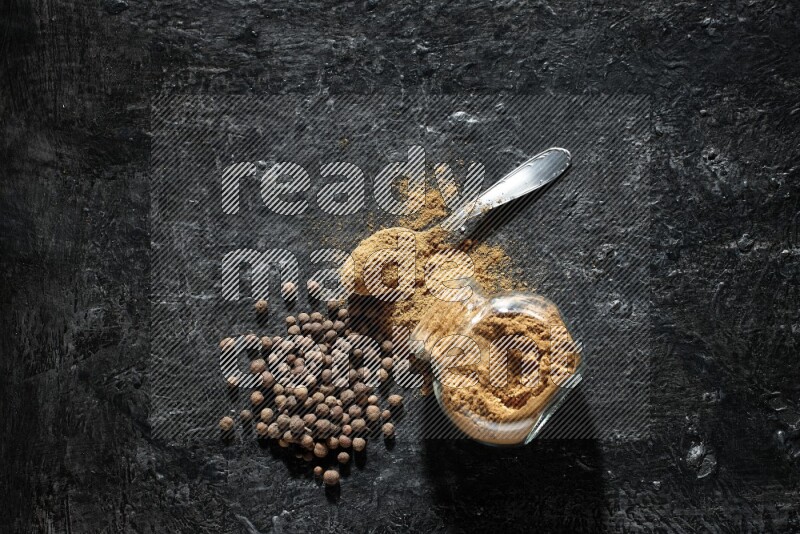 وعاء زجاجي للتوابل مقلوب وملعقة معدنية ممتلئان ببودرة بهار الفلفل الحلو والبودرة تناثرت من الوعاء مع بعض الحبوب على خلفية سوداء