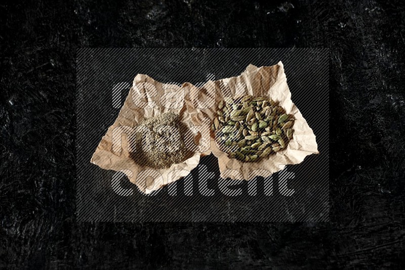 Cardamom seeds and cardamom powder in 2 crumpled pieces of paper on textured black flooring