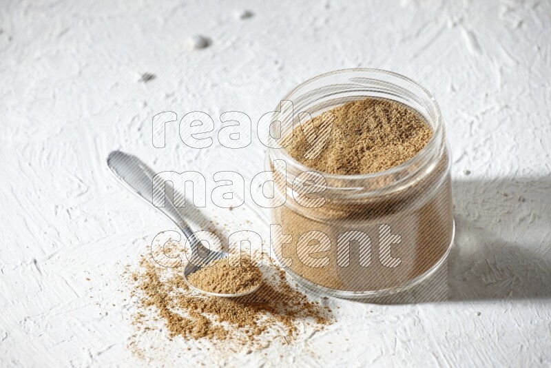 A glass jar and a metal spoon full of cumin powder on textured white flooring
