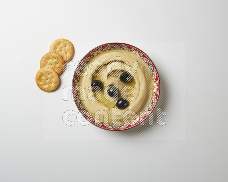 Hummus in a red plate with patterns garnished with black olives on a white background