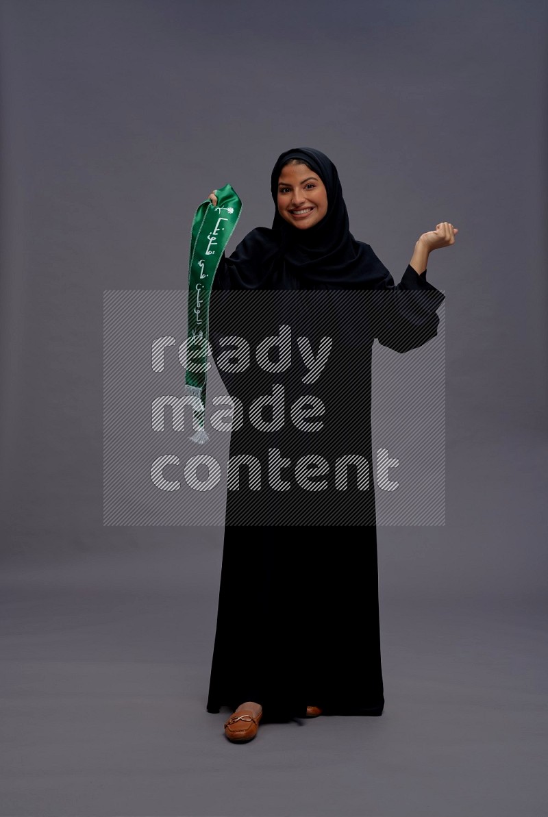 Saudi woman wearing Abaya standing holding Saudi flag on gray background