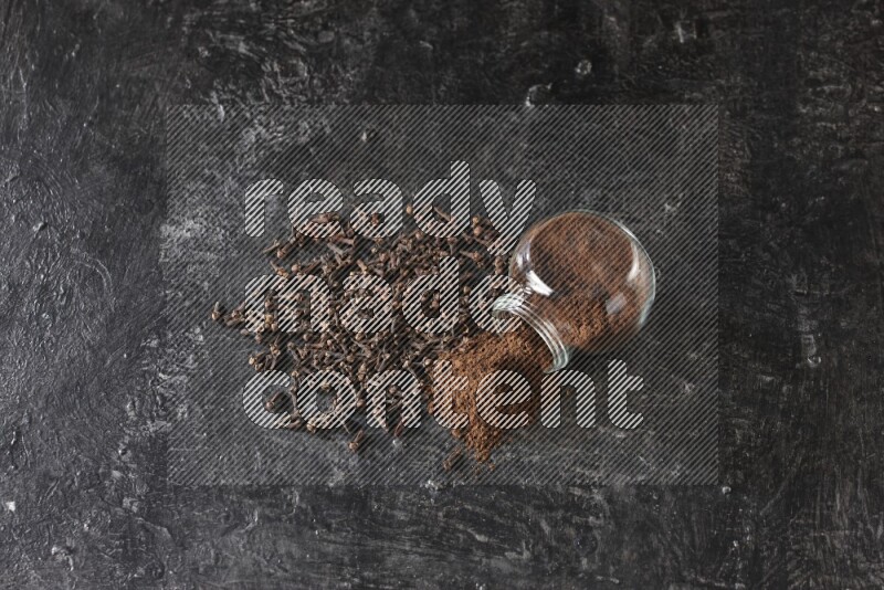 وعاء زجاجي للبهارات مقلوب ممتلئ ببودرة القرنفل والبودرة سقطت منه مع حبوب القرنفل المتناثرة على أرضية سوداء