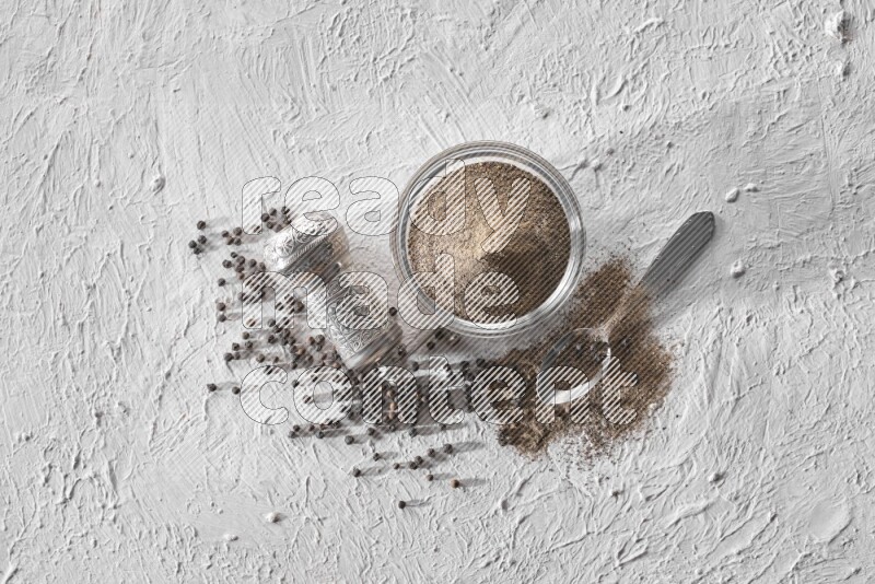 فلفل أسود مطحون مع حبوب الفلفل الأسود ومطحنة معدنية وملعقة معدنية علي خلفية بيضاء