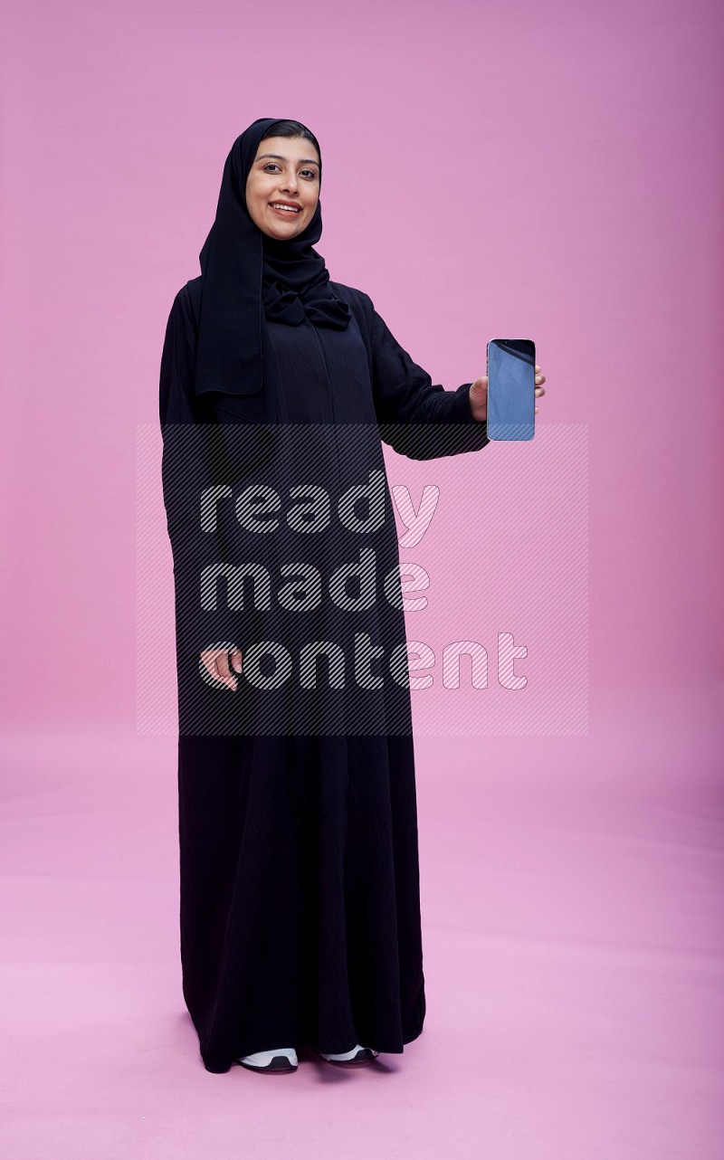 Saudi woman wearing Abaya standing showing phone to camera on pink background