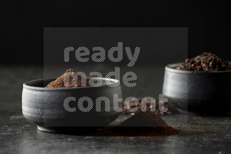 2 Black pottery bowls full of cloves and the other full of cloves powder on textured black flooring