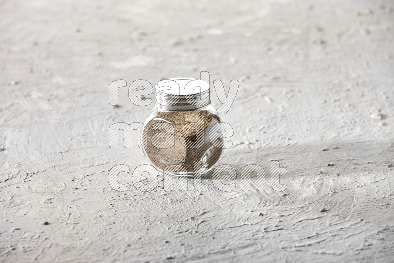 A glass spice jar full of powder black pepper on textured white flooring