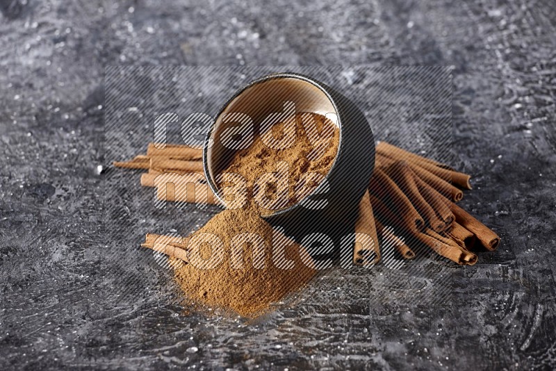 Black pottery bowl over filled with cinnamon powder and cinnamon sticks around the bowl on a textured black background