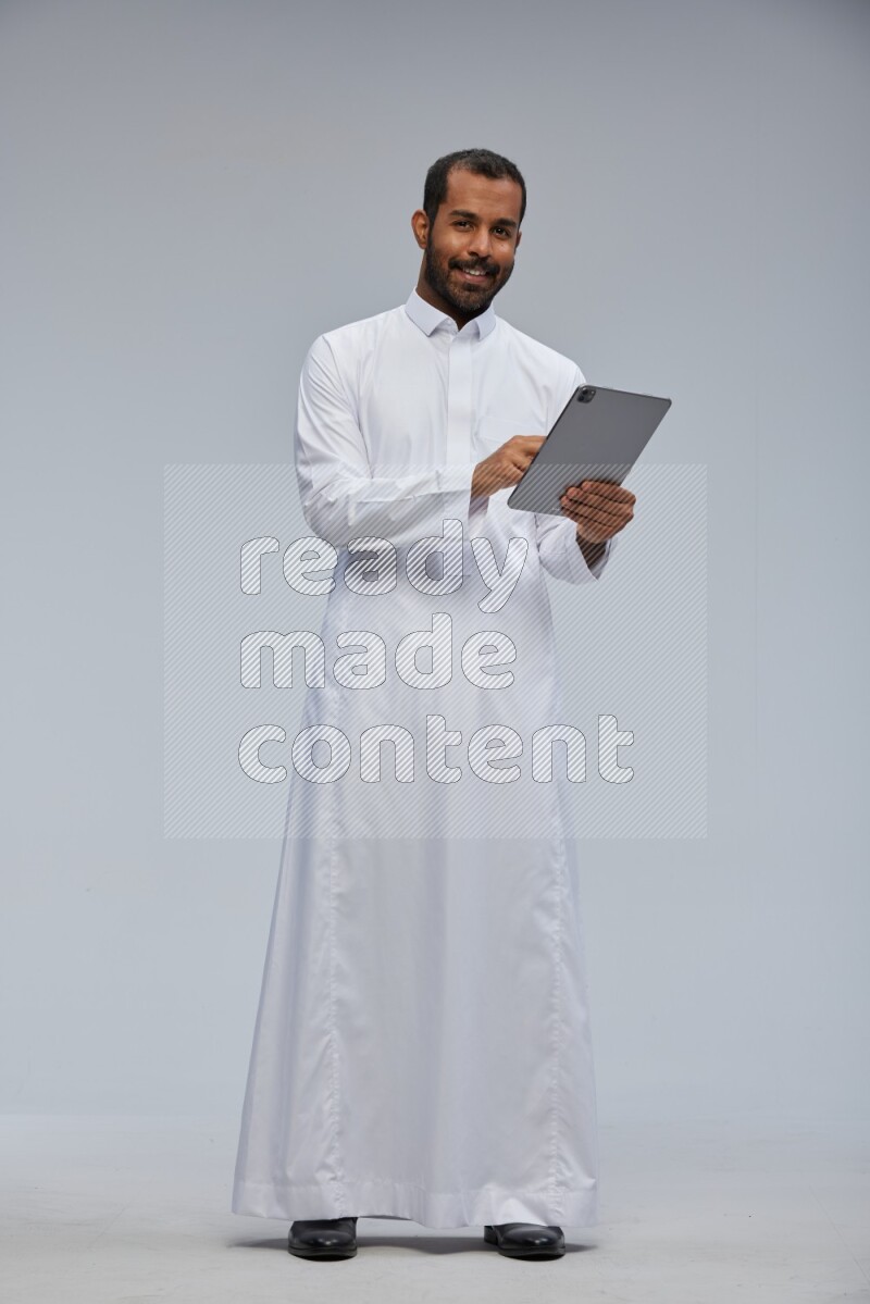 Saudi man wearing Thob standing working on tablet on Gray background