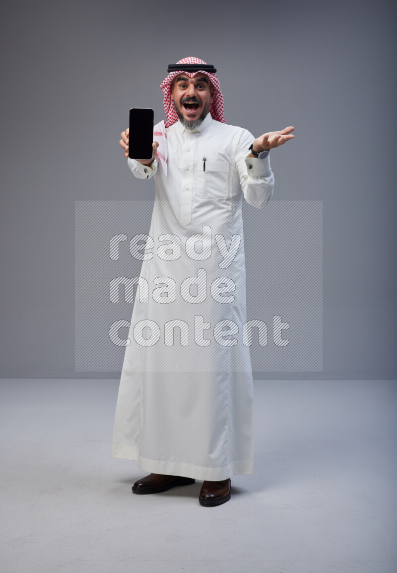 Saudi man Wearing Thob and red Shomag standing showing phone to camera on Gray background