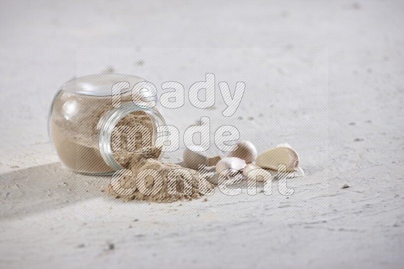 A glass spice jar full of garlic powder flipped over with the powder came out on a textured white flooring