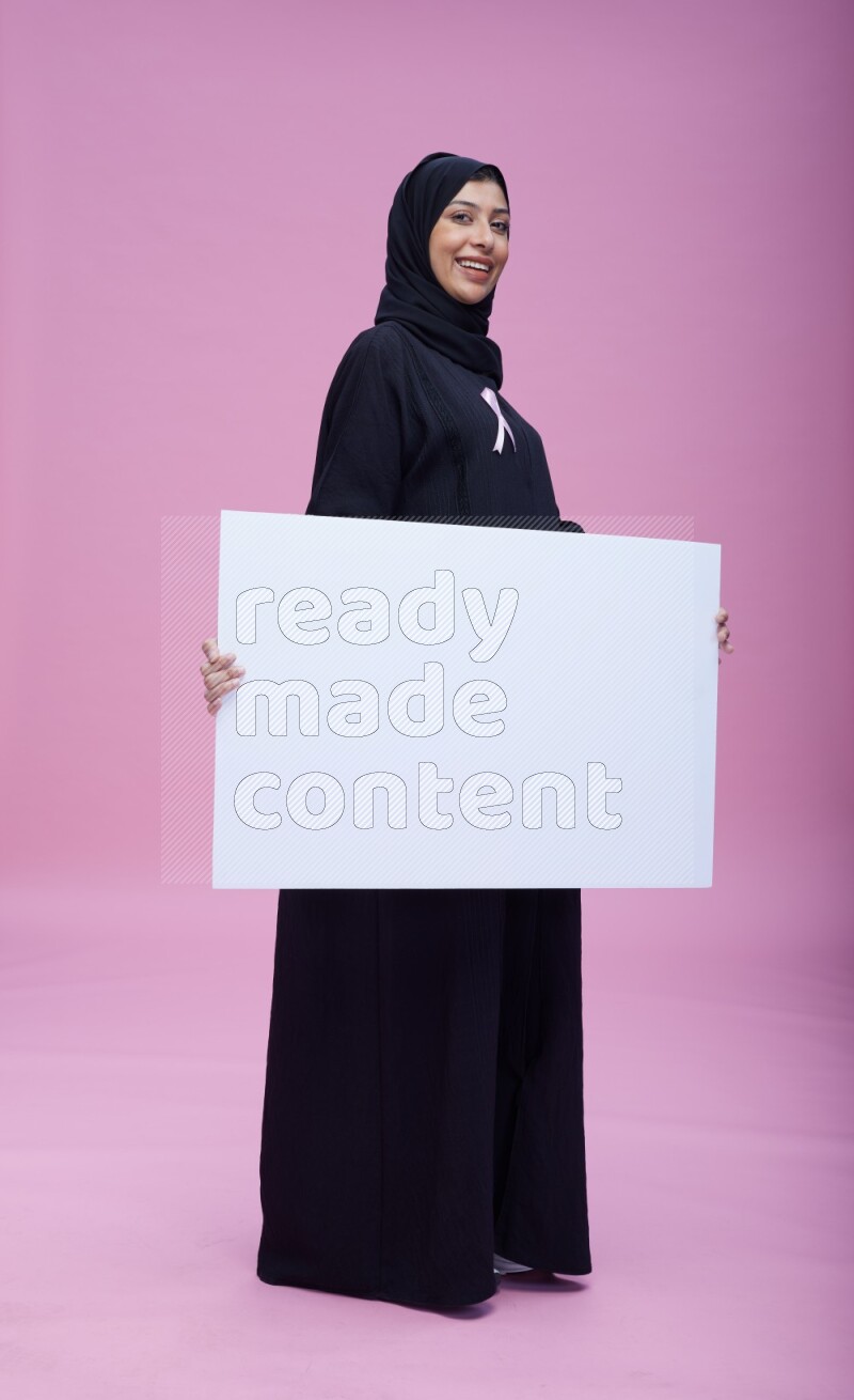Saudi woman wearing pink ribbon on Abaya standing holding board on pink background