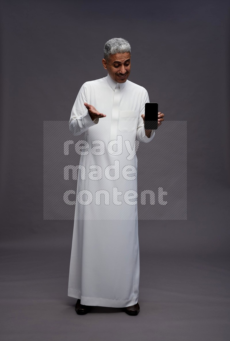 Saudi man wearing thob standing showing phone to camera on gray background