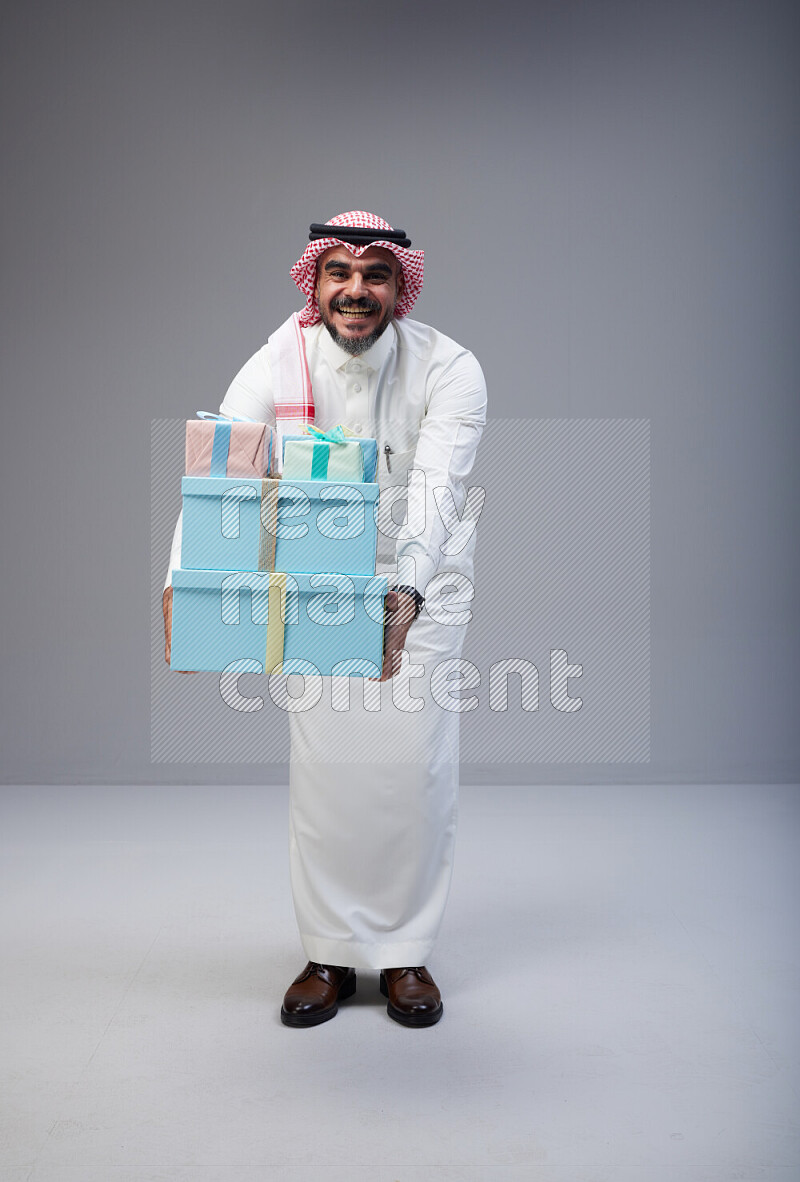 Saudi man Wearing Thob and red Shomag standing holding gift box on Gray background