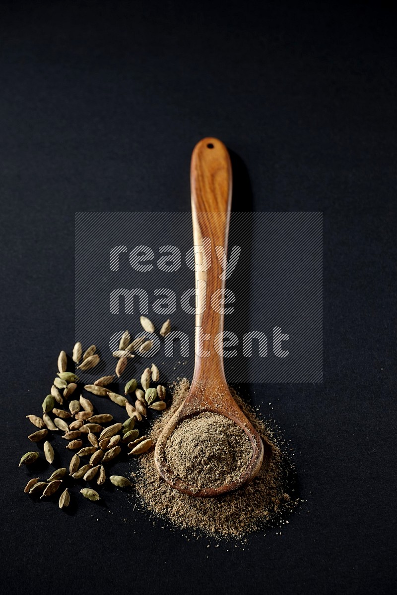 A wooden ladle full of cardamom powder and cardamom seeds beside it on black flooring