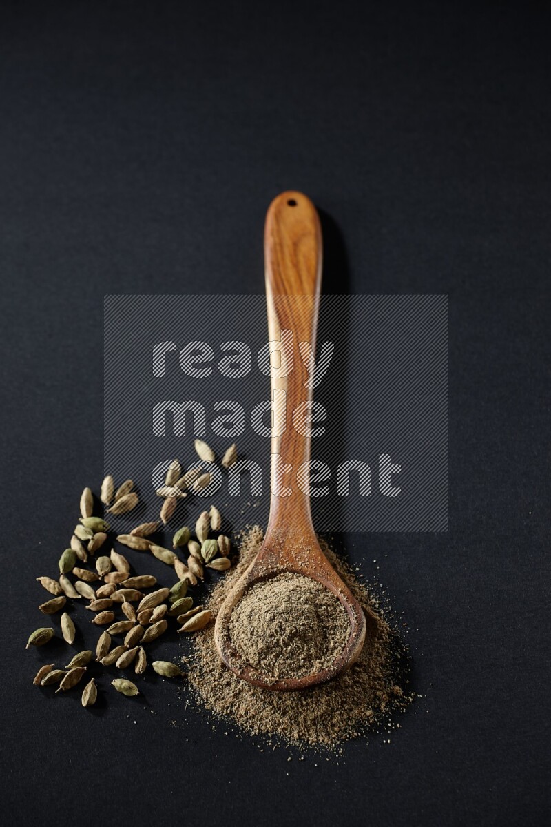 A wooden ladle full of cardamom powder and cardamom seeds beside it on black flooring