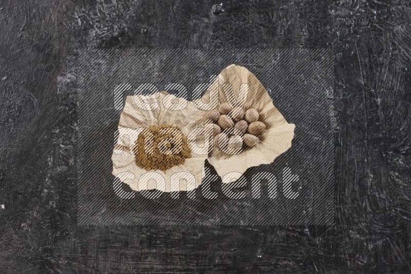 2 crumpled piece of paper full of nutmeg seeds and powder on a textured black flooring
