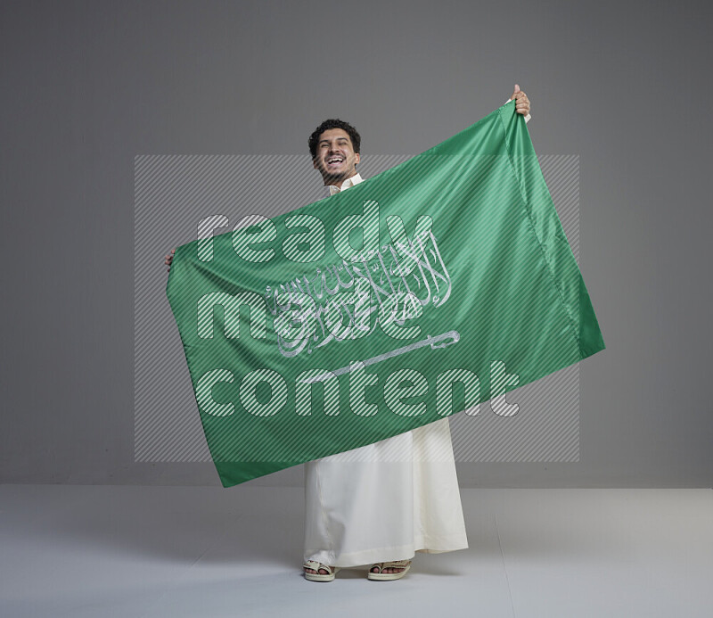 A Saudi man standing wearing thob holding big Saudi flag on gray background