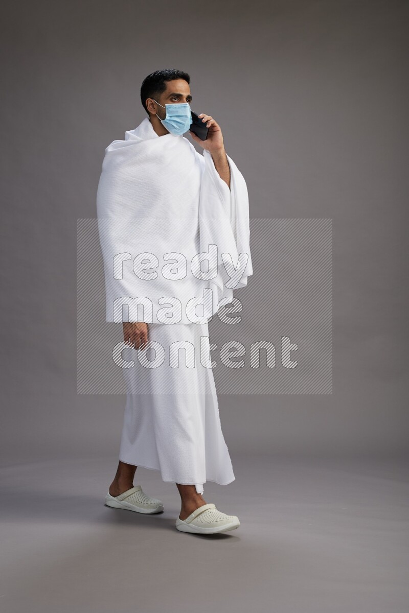 A man wearing Ehram with face mask Standing talking on phone on gray background