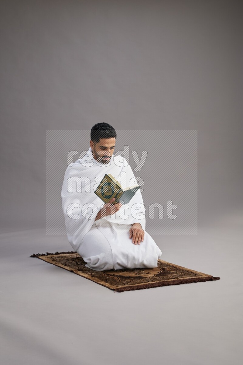 A man wearing Ehram sitting on floor reading quran on gray background