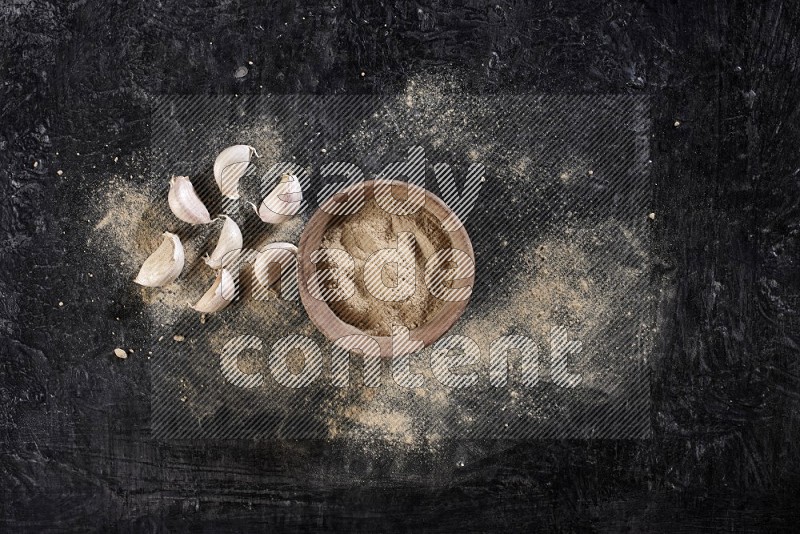 A wooden bowl full of garlic powder and beside it garlic cloves on a textured black flooring in different angles