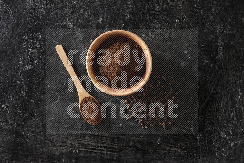 وعاء خشبي وملعقة خشبية ممتلئان ببودرة القرنفل مع حبوب القرنفل المتناثرة على خلفية سوداء