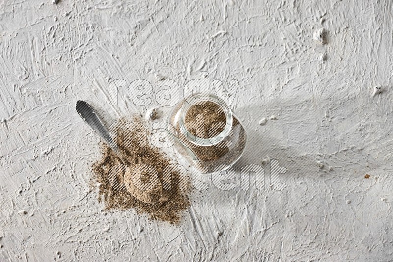 A glass spice jar full of black pepper powder and metal spoon full of it on textured white flooring