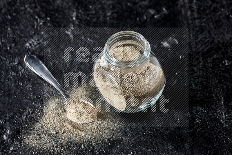 Herbal glass jar and metal spoon full of white pepper powder on textured black flooring