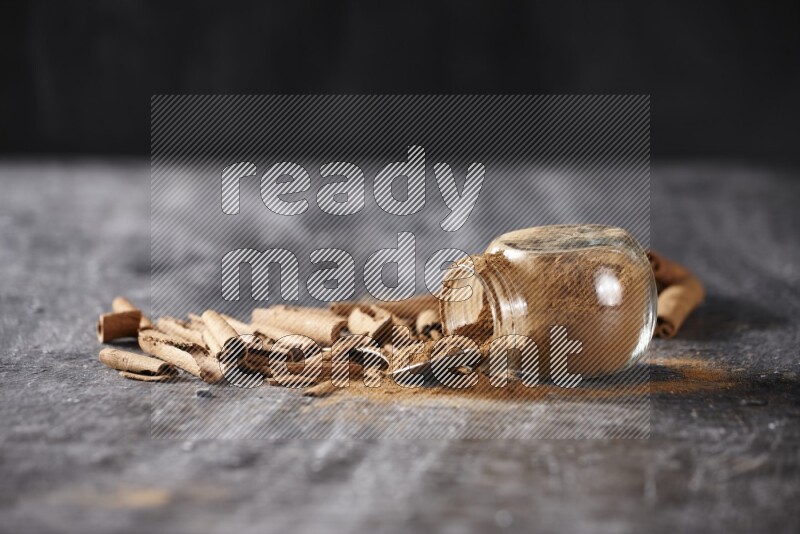 Herbal glass jar full cinnamon powder flipped and a metal spoon full of powder surrounded by cinnamon sticks on textured black background in different angles