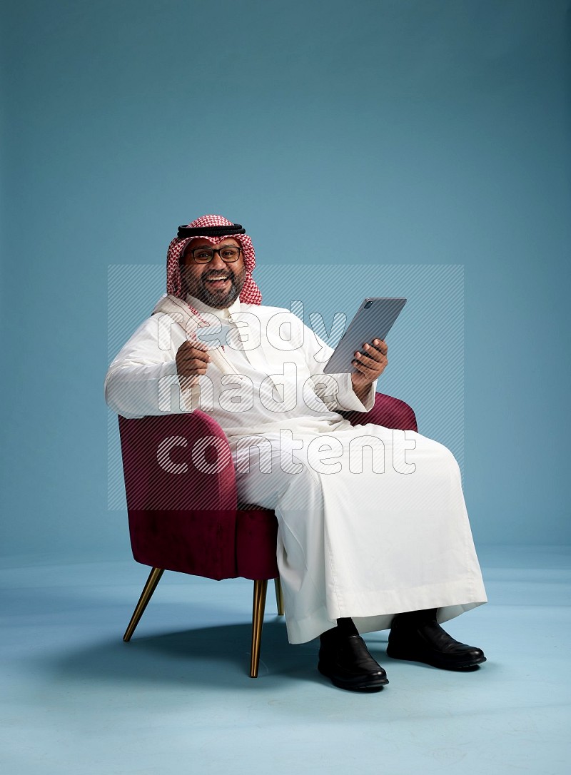 Saudi Man with shimag sitting on chair holding ATM card while working on tablet on blue background